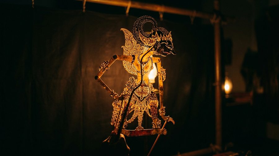 Wayang kulit berukir motif emas disinari cahaya dari belakang dalam ruangan gelap